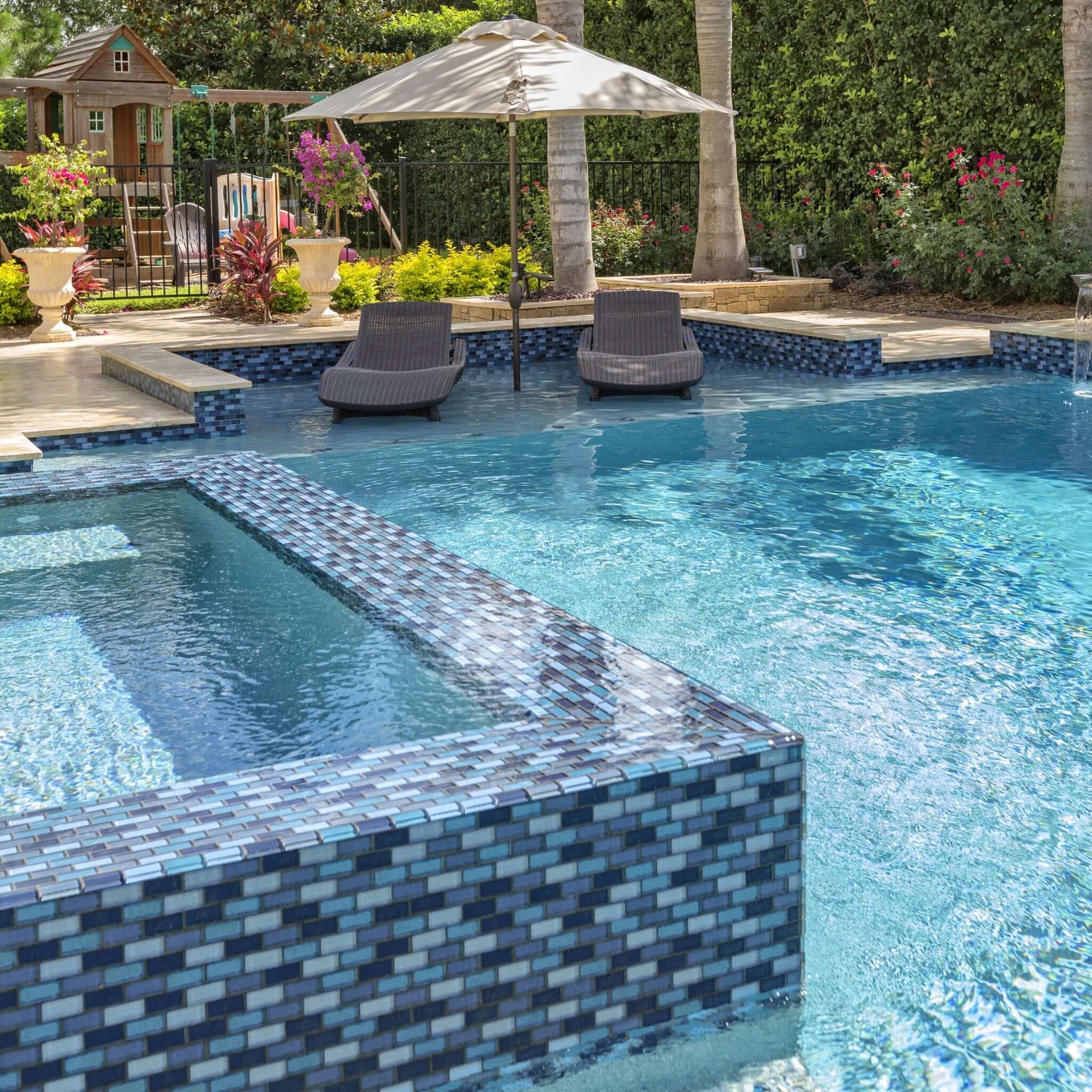 Relaxing pool area with aqua blend glass tiles, featuring lounge chairs and an umbrella in a vibrant outdoor setting.