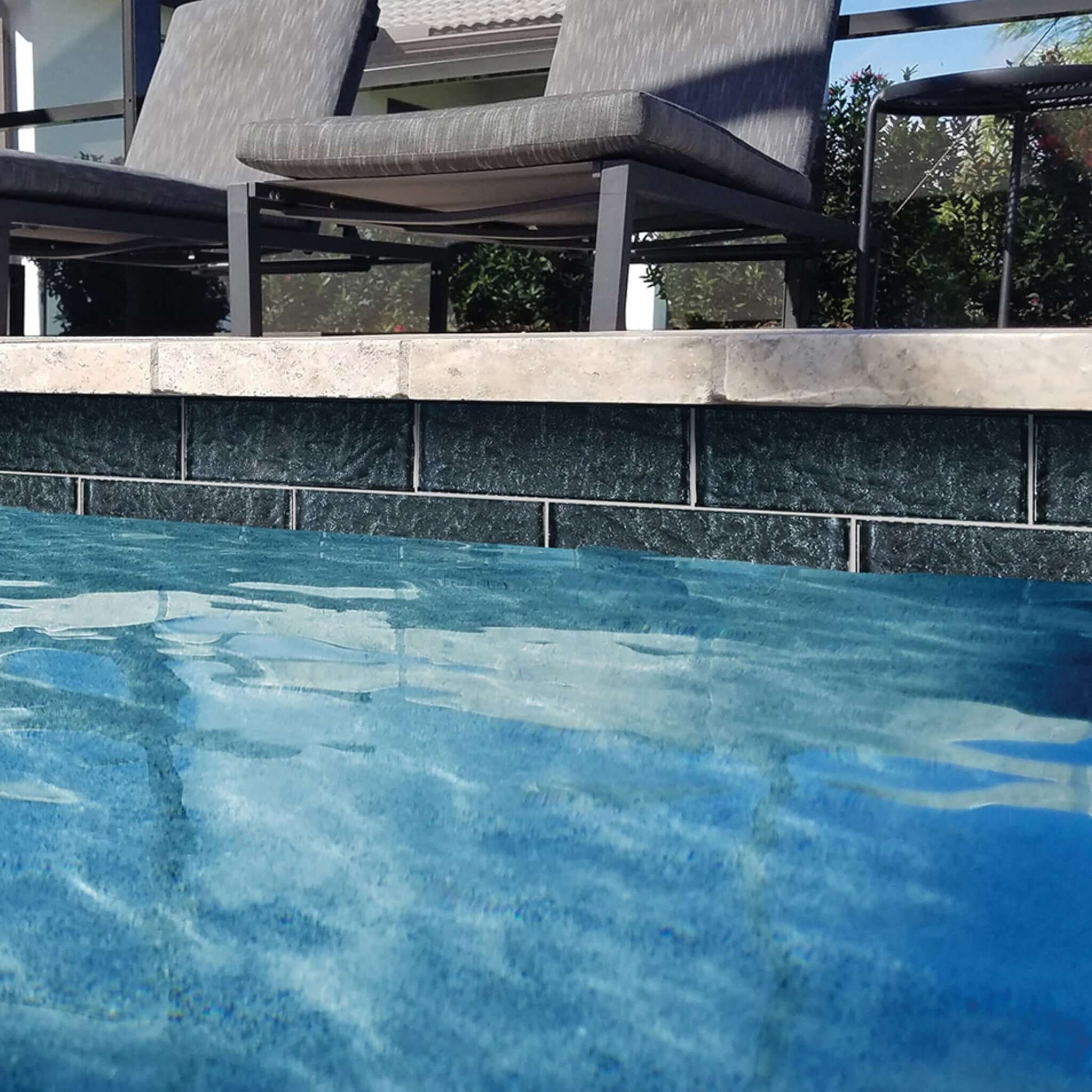 Sparkling blue swimming pool water with sleek black tiles and modern lounge chairs set against a sunny backdrop.