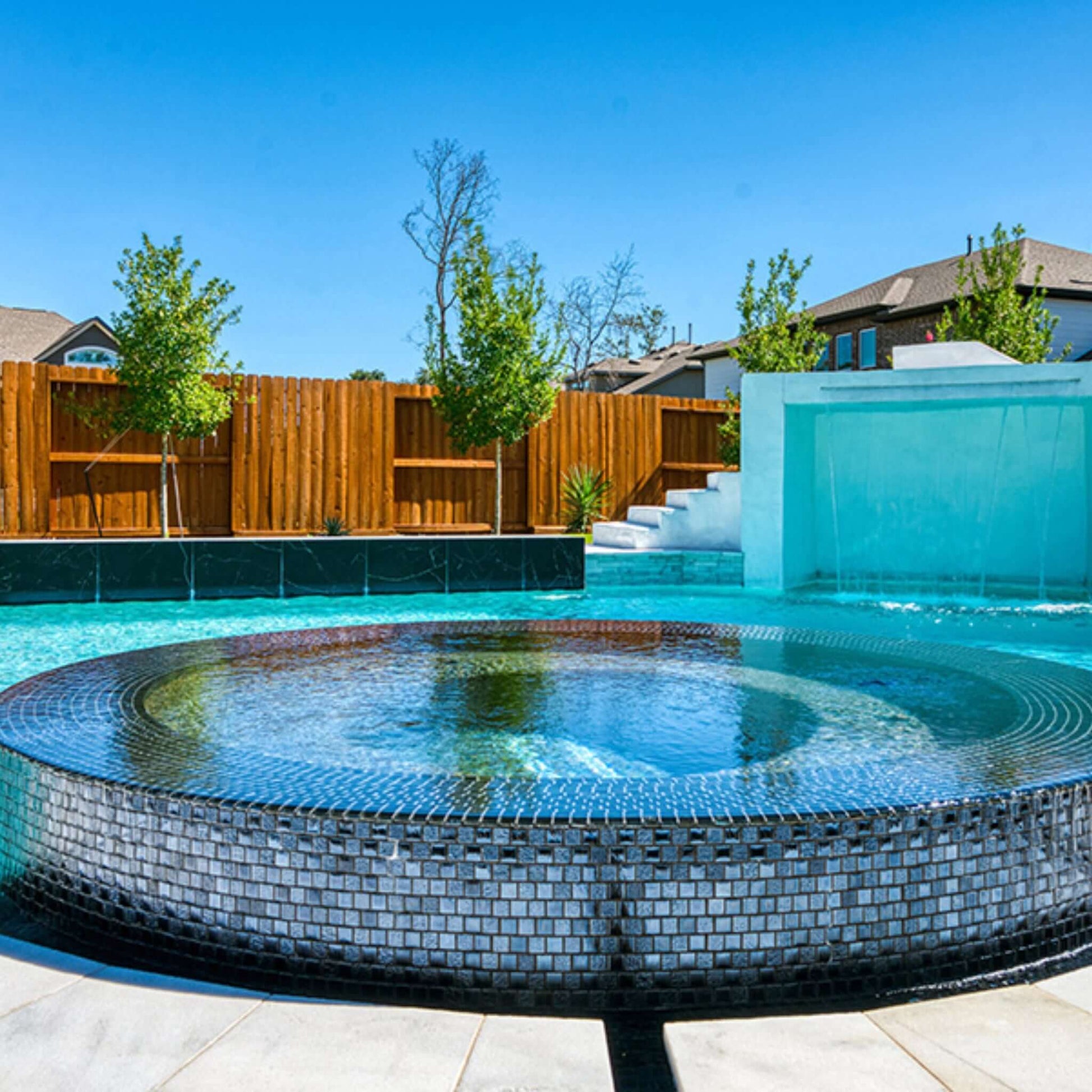 Modern pool with circular mosaic design and water feature, surrounded by trees and wooden fence. Perfect for relaxation.