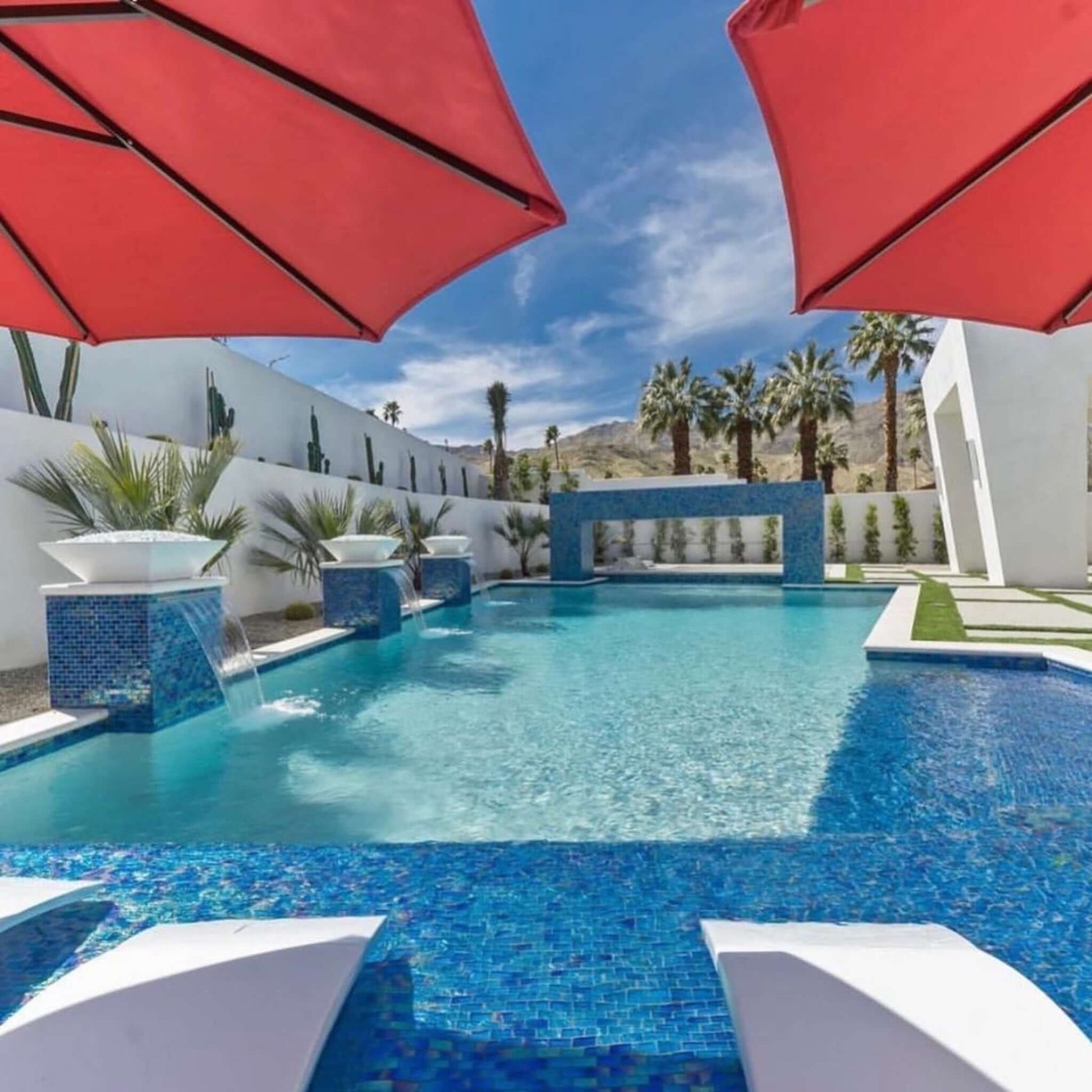 Luxury outdoor pool area with red umbrellas, mosaic tiles, and palm trees against a desert backdrop.